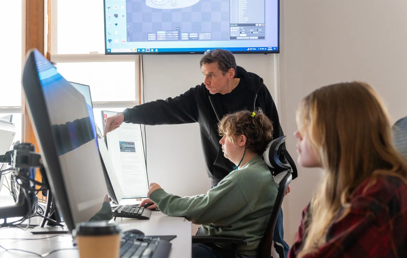 Instructor pointing at a monitor while two students work at computers in a classroom.