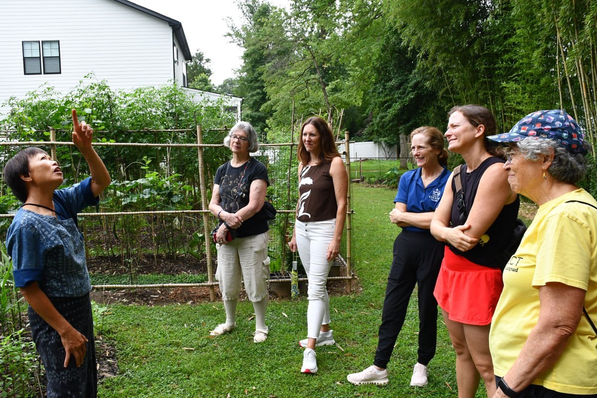 A woman gestures upward while giving a garden tour to a small group of visitors in a backyard with vegetable plants and bamboo stakes.