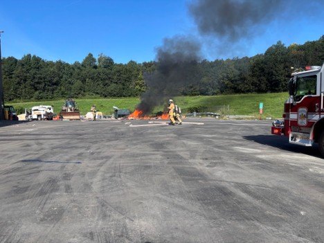 Firefighters in turnout gear approach a fire with black smoke rising at an open paved facility.