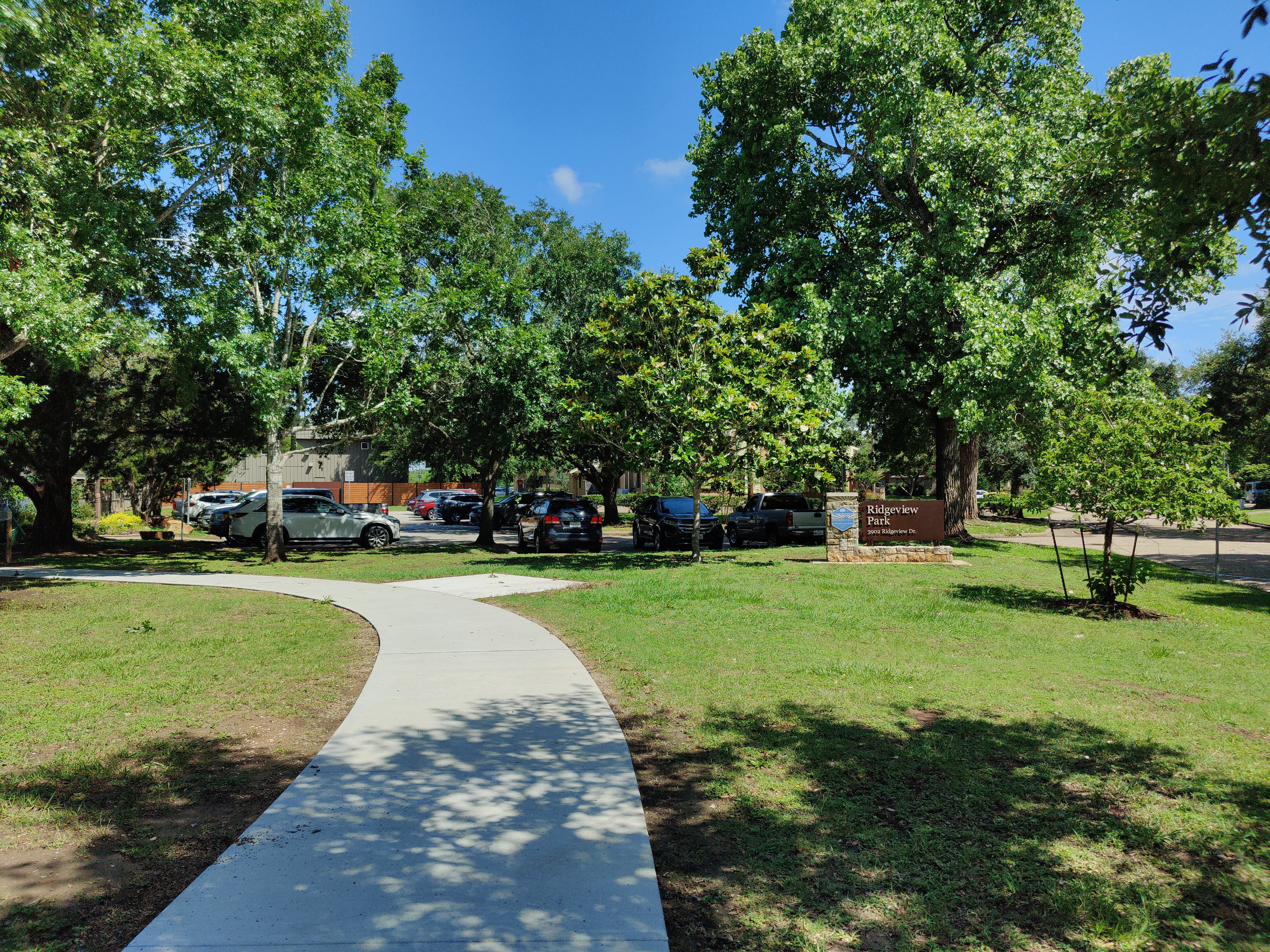 Curved paved path through a green park with large shade trees and a sign.