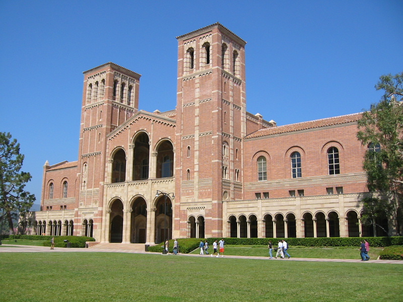 Red brick university building with twin towers and arched colonnades on a sunny day.