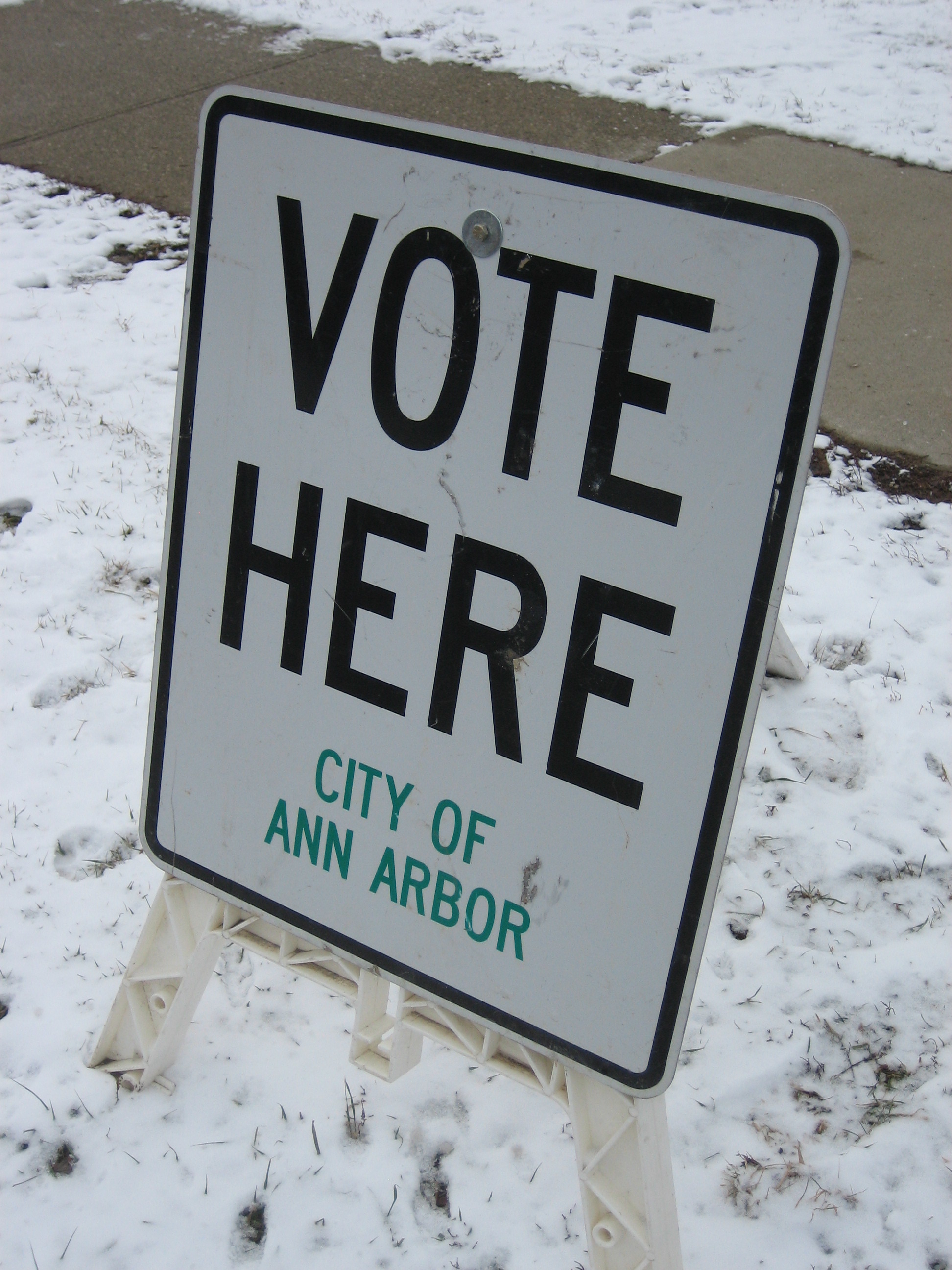 A portable white sign reading "Vote Here" stands on snow-covered ground outside a polling place.