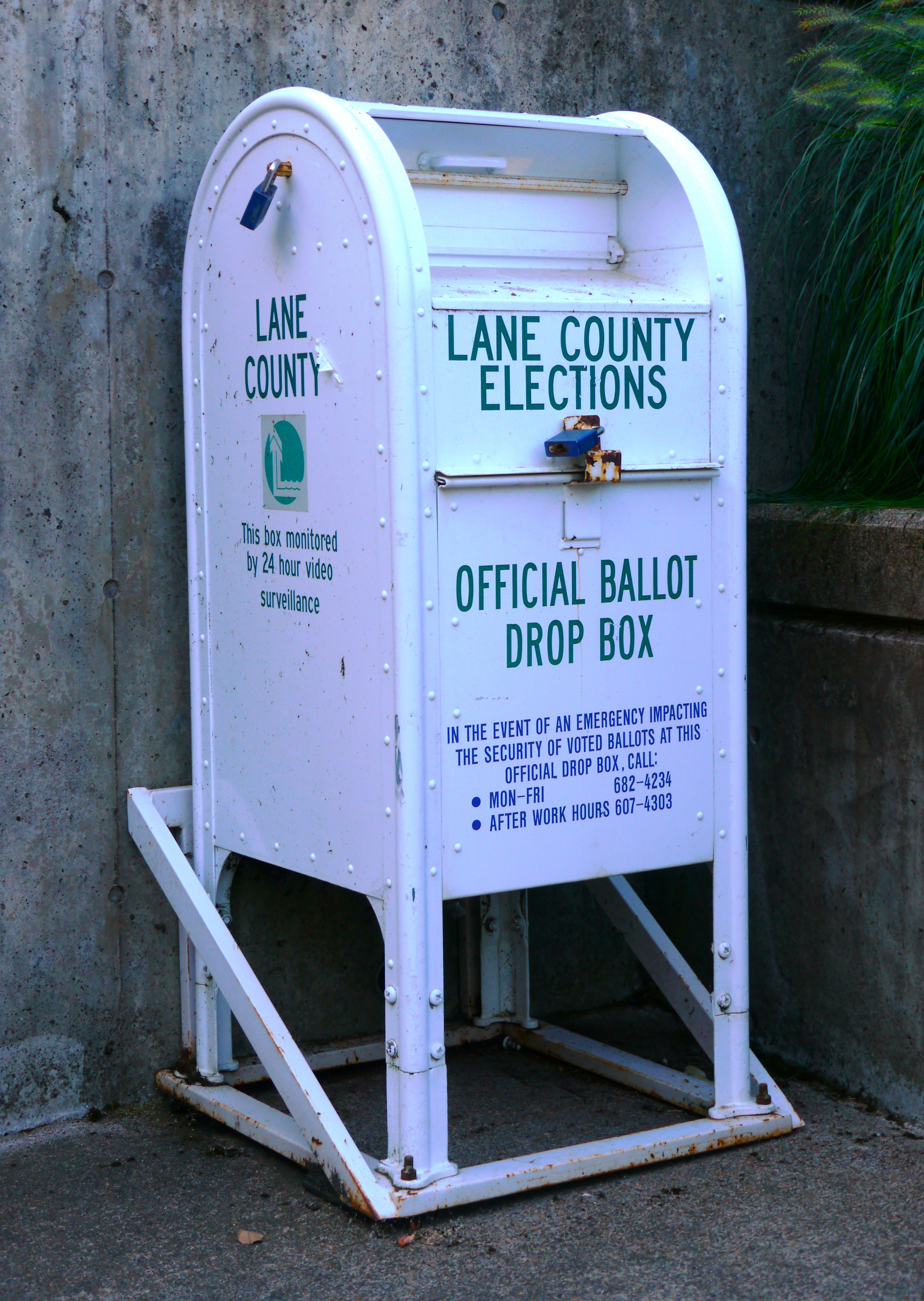 A white metal official ballot drop box mounted on a metal stand outdoors.