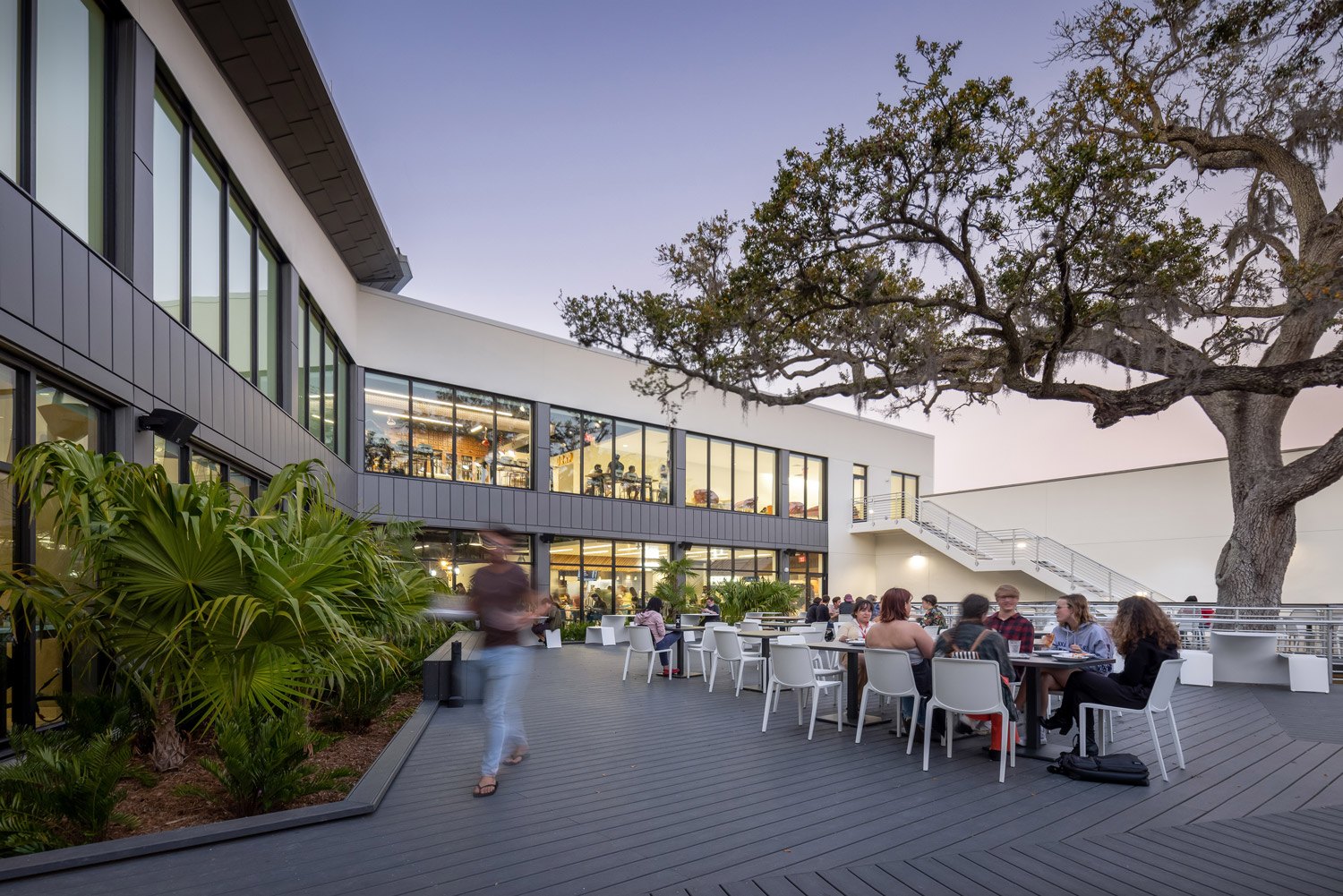 Modern two-story academic building with outdoor wooden deck and tropical landscaping at dusk.