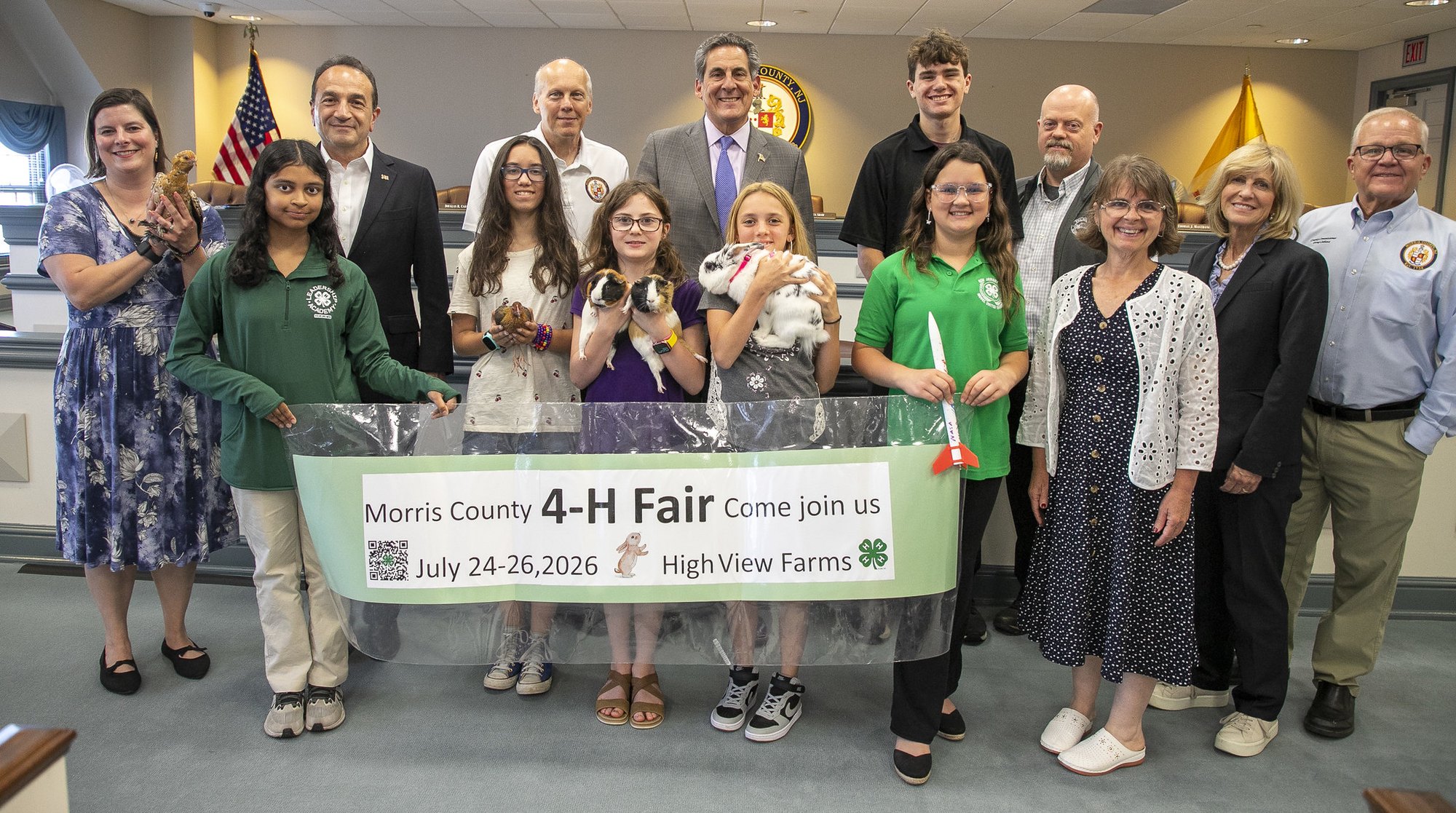 Children in green shirts hold small animals while posing with adults indoors near American and state flags.