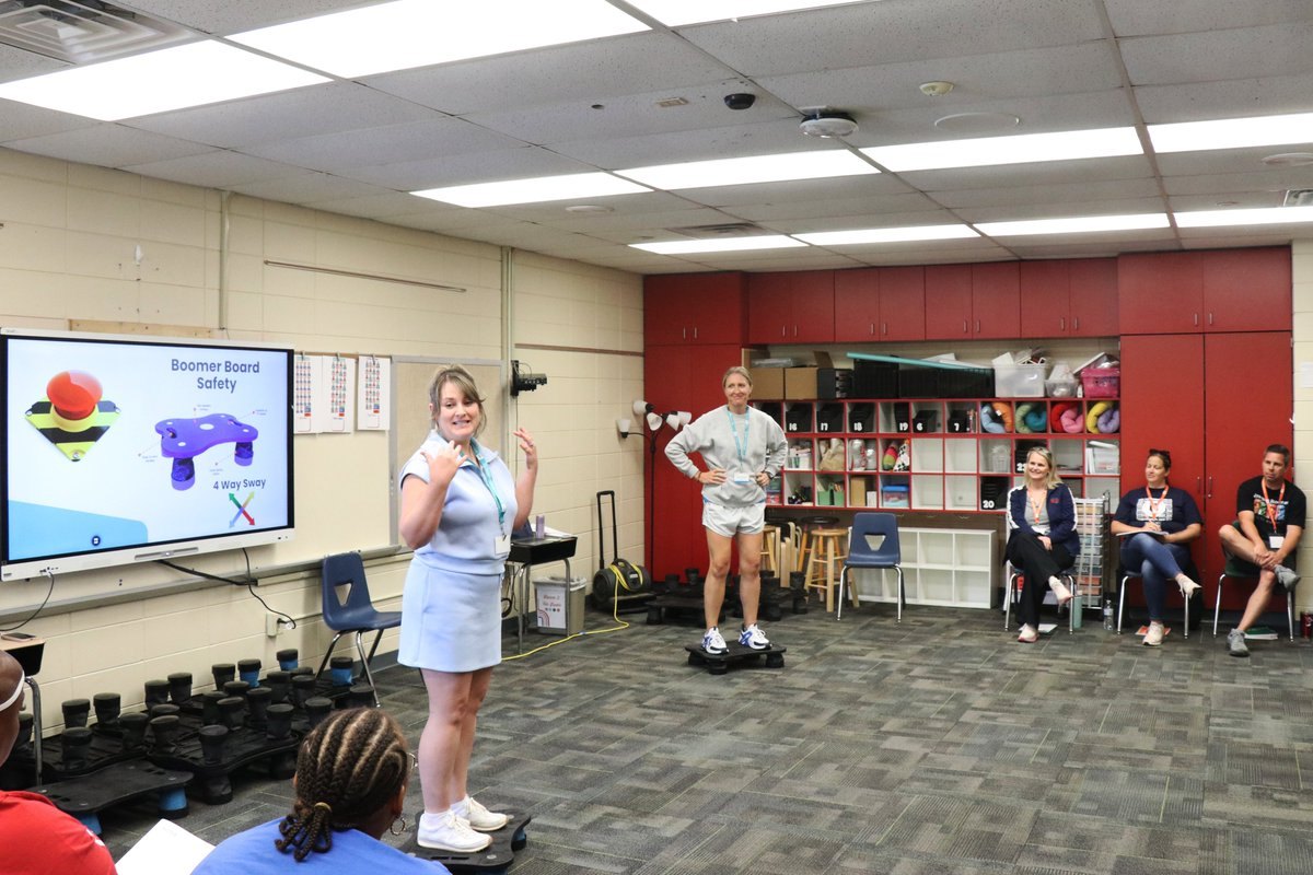 A woman stands on a balance board presenting to a group of seated educators in a classroom.