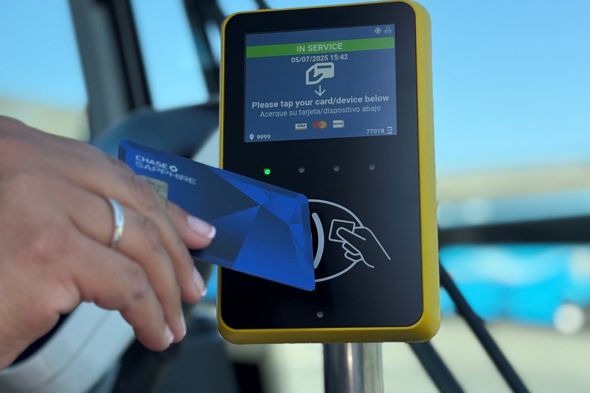A hand tapping a credit card on a yellow contactless payment reader inside a bus.
