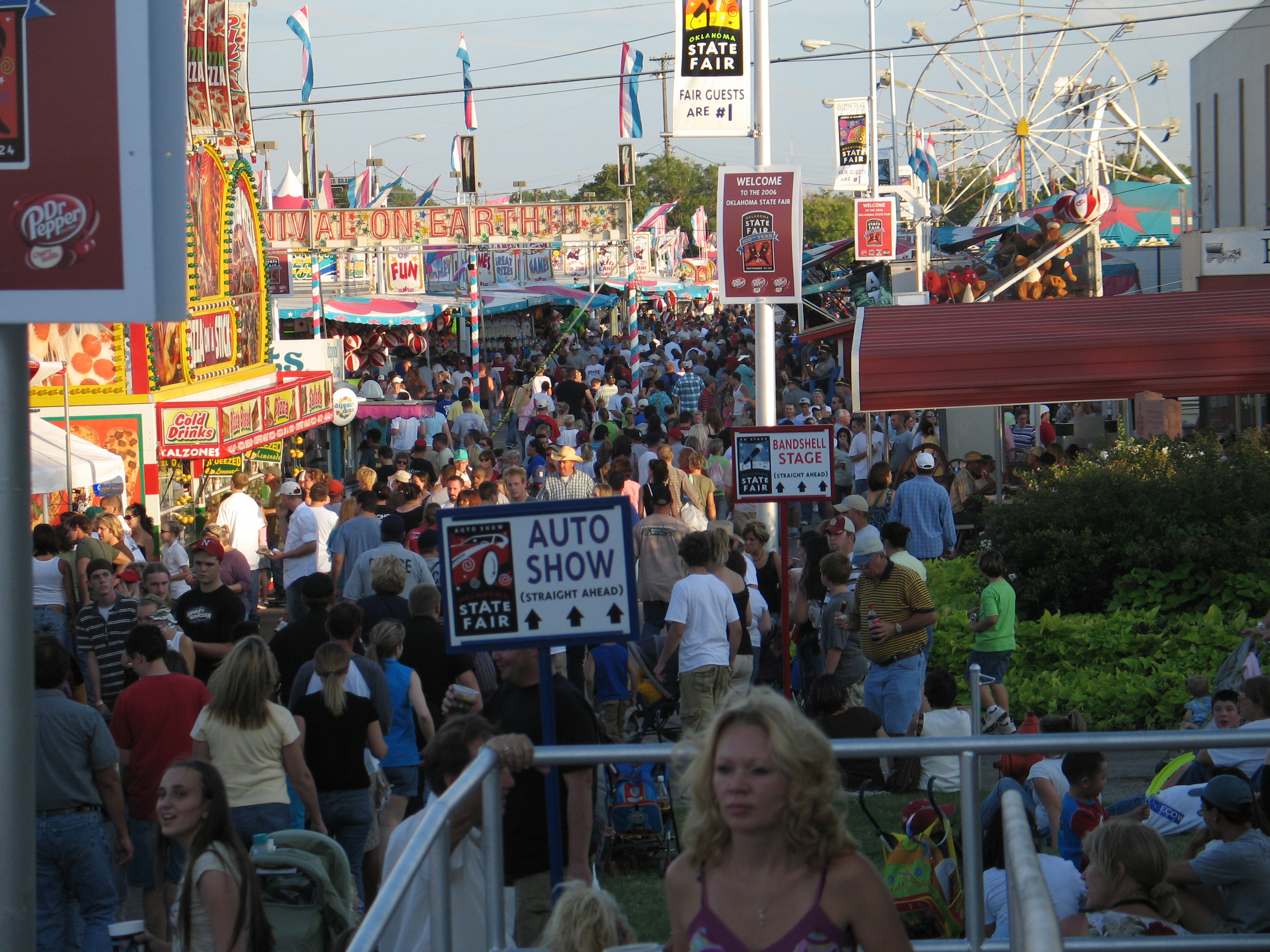 Crowds walk through a busy fairground midway with a Ferris wheel in the background.