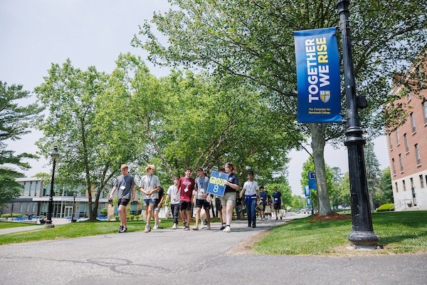 Students walk along a tree-lined campus path past a lamp post with a blue banner.