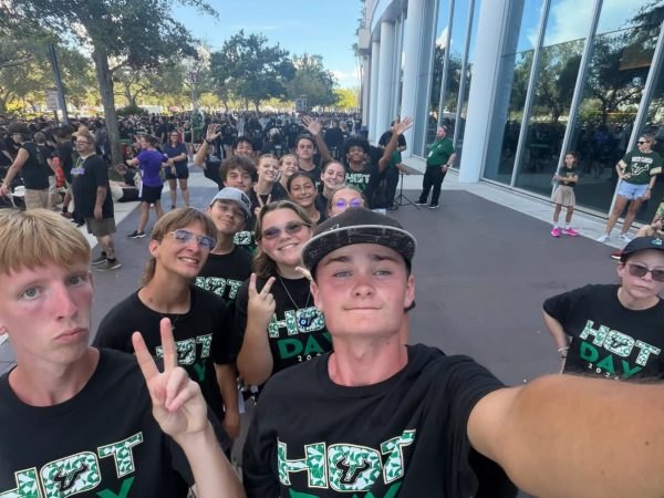 Teenagers in matching black HOT Day T-shirts pose for a selfie outside a stadium building.