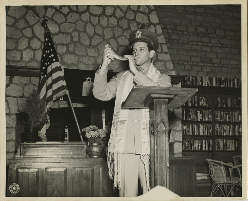 A uniformed chaplain wearing a prayer shawl blows a shofar at a wooden podium.