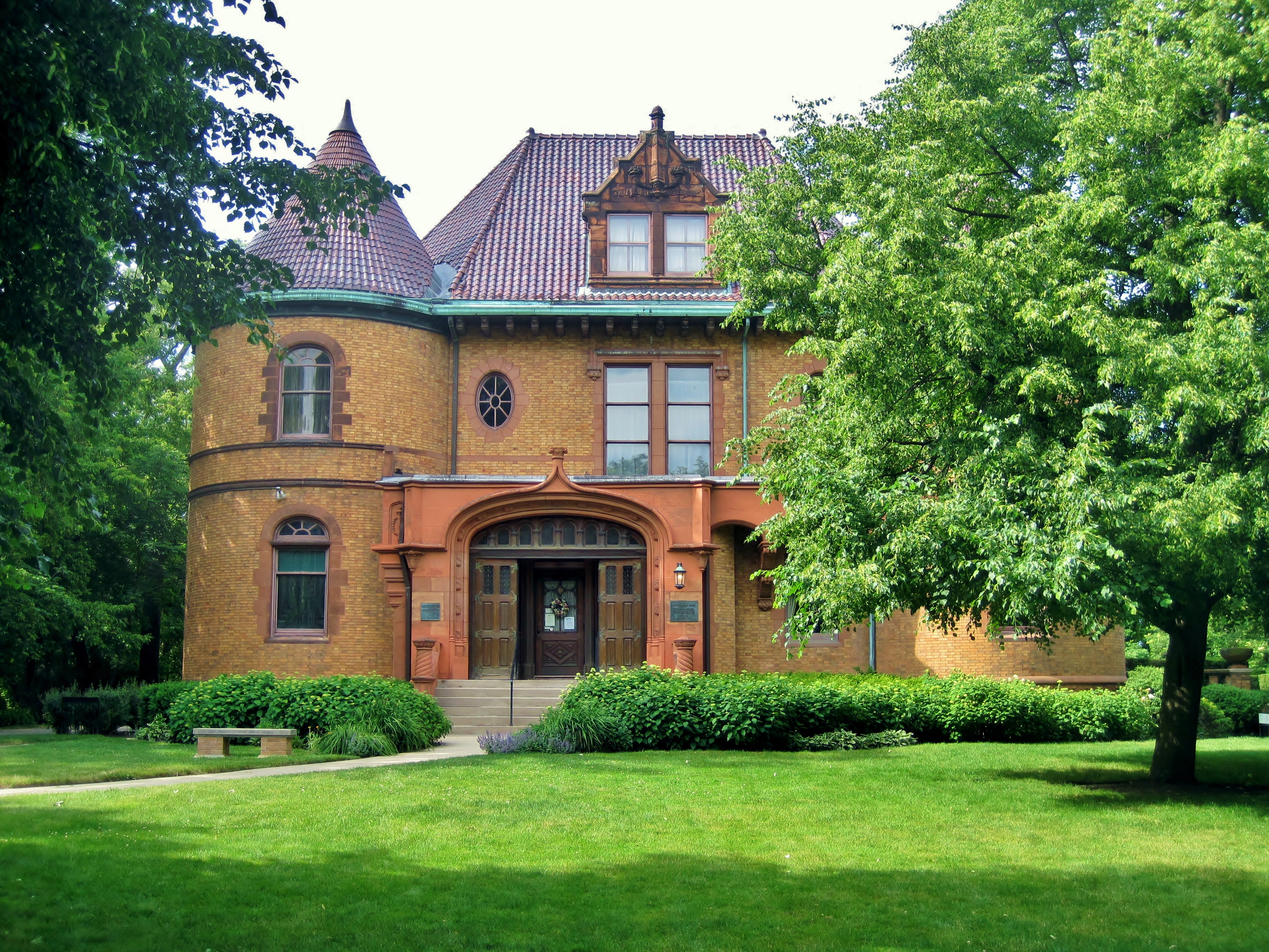 Brick chateauesque mansion with red tile roof and turret, surrounded by mature trees.