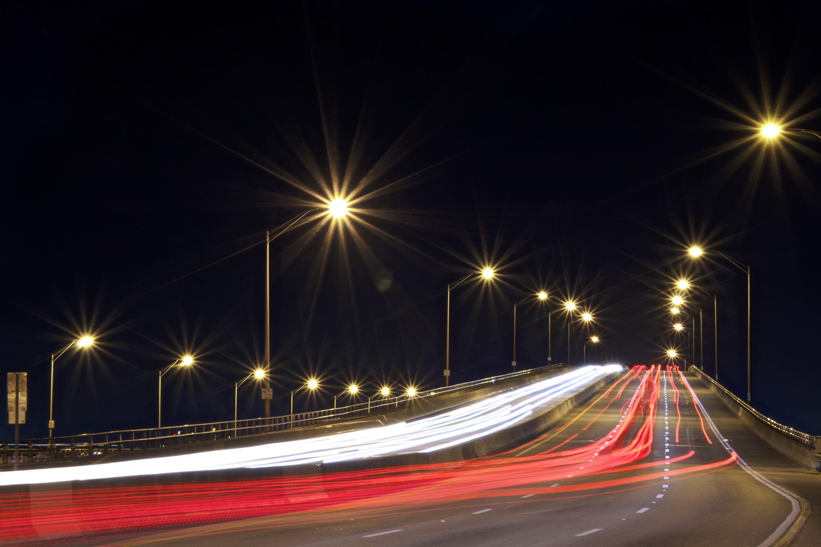 Rows of bright street lamps line a bridge at night with vehicle light trails below.