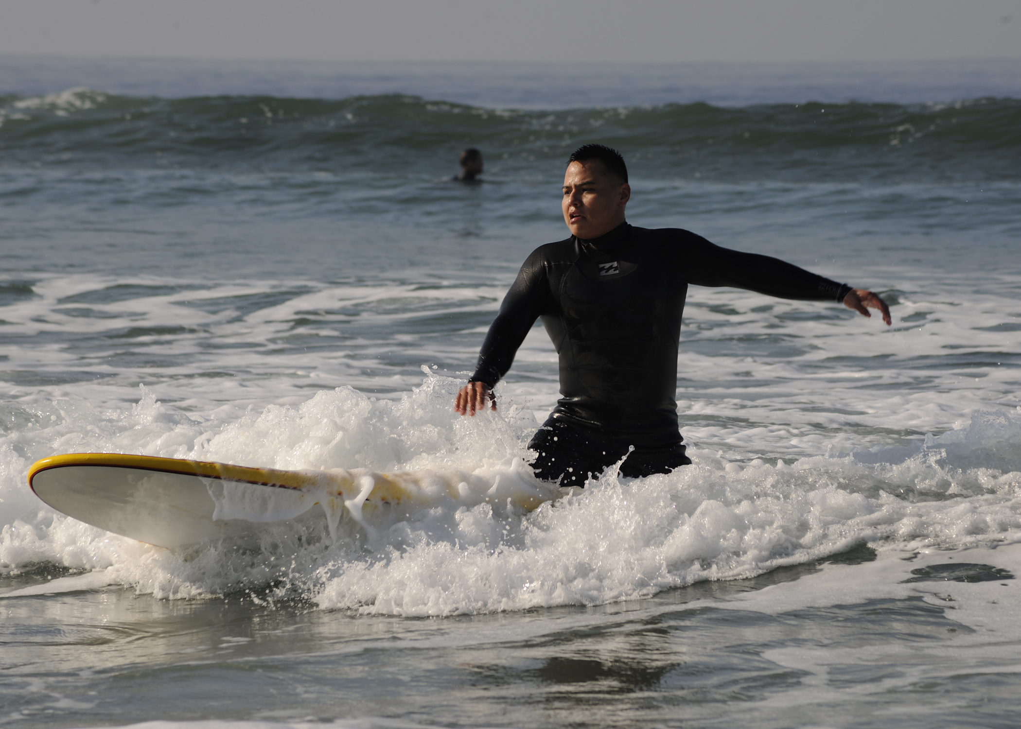 Surfer in black wetsuit riding a yellow-and-white surfboard through shallow ocean waves.
