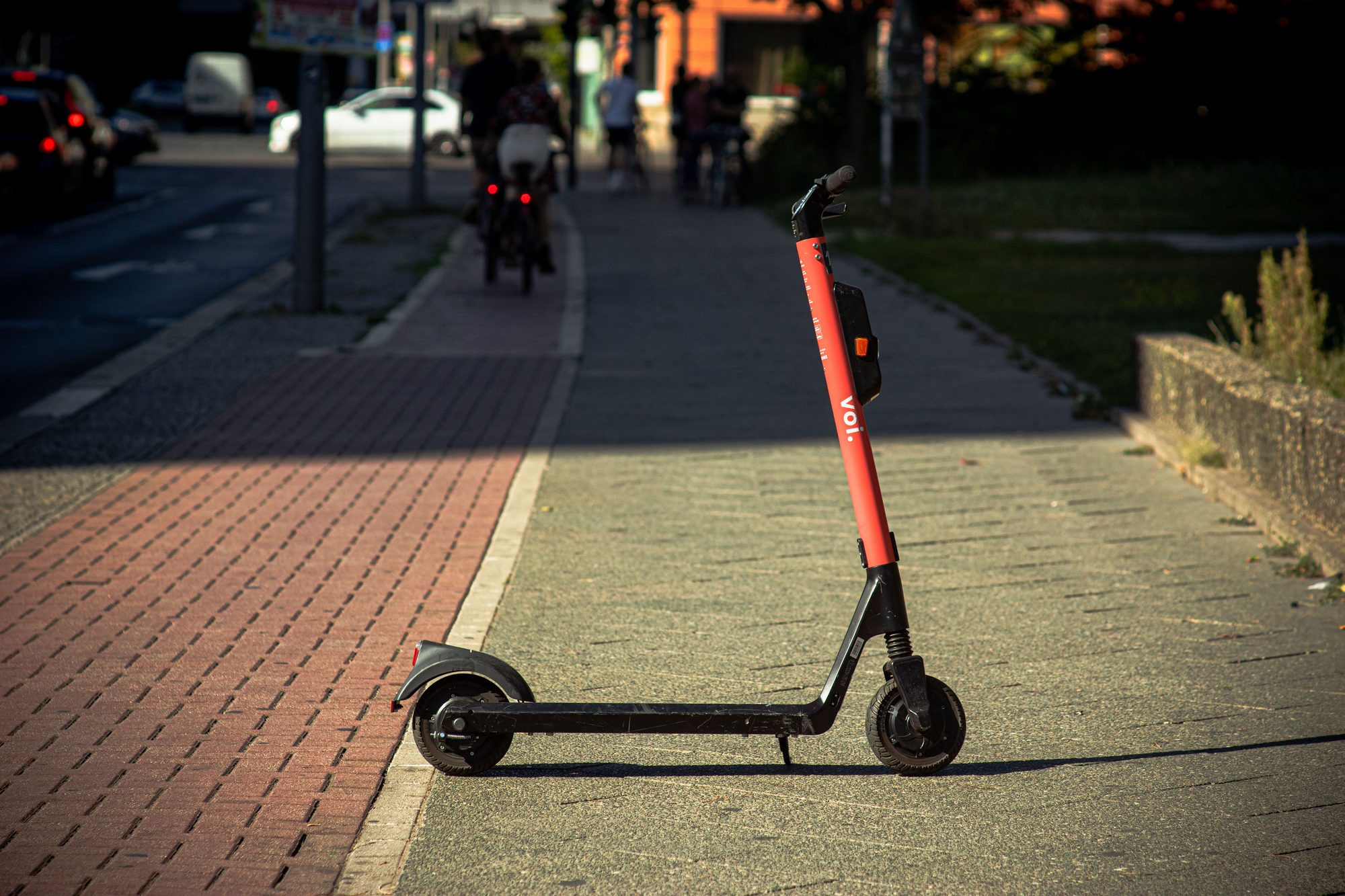 A Voi e-scooter parked on a sunlit city sidewalk with a blurred urban background.