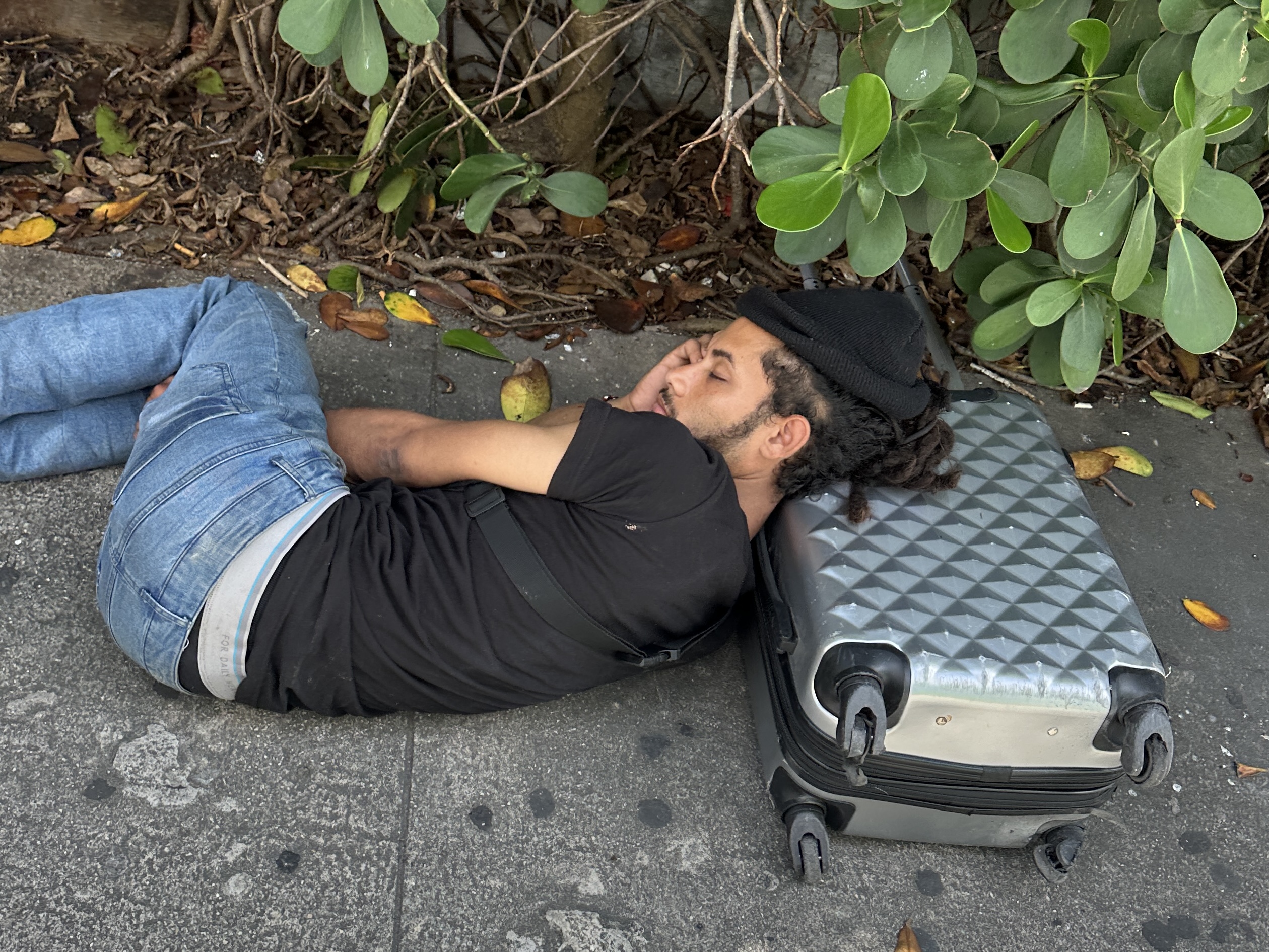 A man lies on a concrete sidewalk using a rolling suitcase as a pillow, with tropical plants behind him.