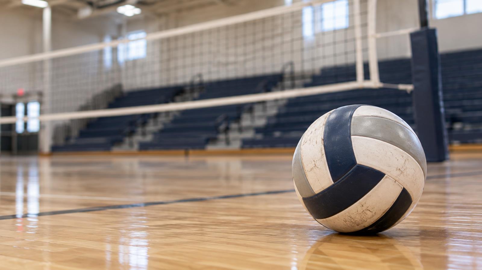 A volleyball resting on a hardwood gymnasium floor near a net with bleachers in the background.