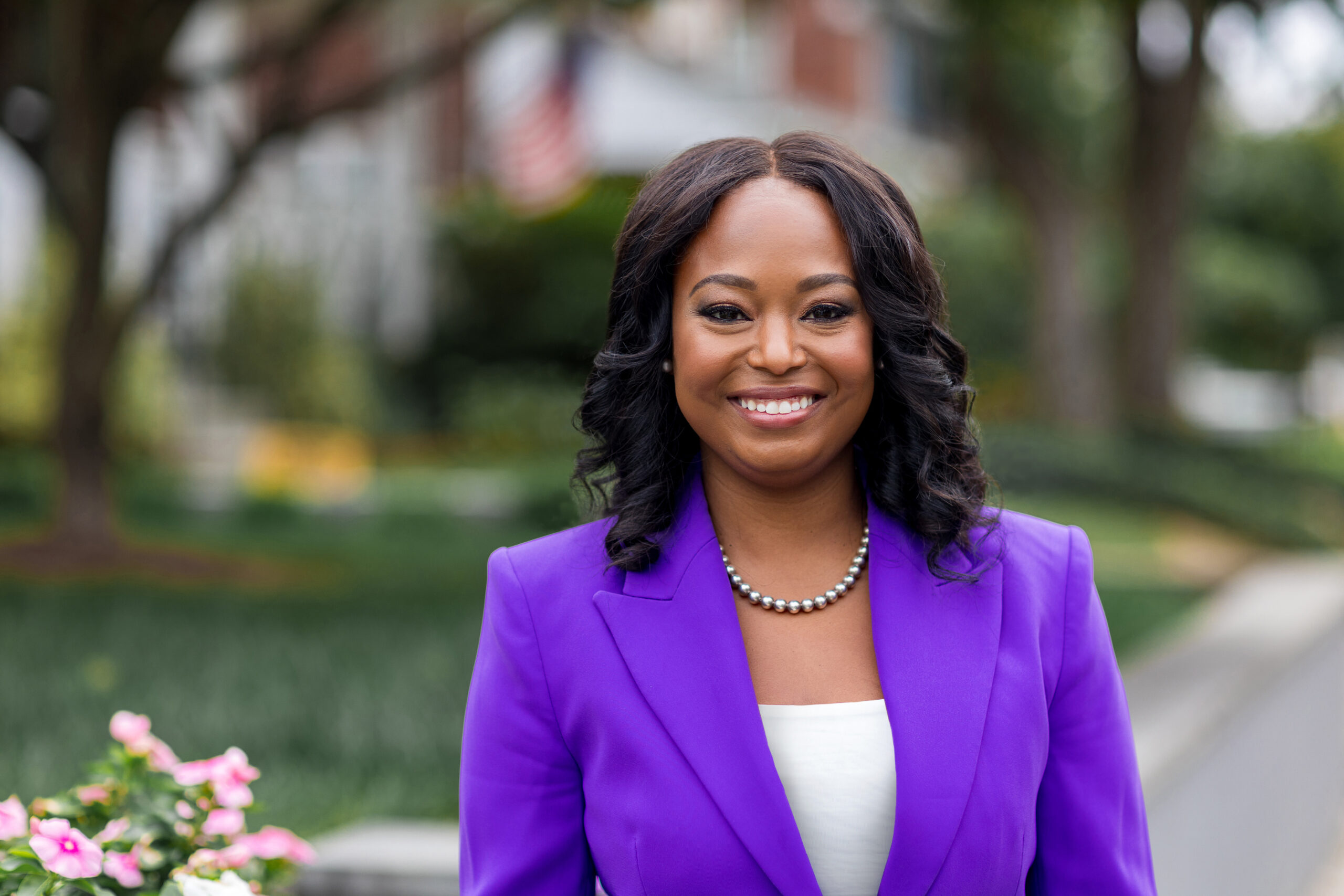 Woman in a purple blazer and pearl necklace smiling outdoors with an American flag in the background.