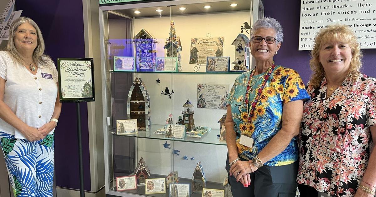 Three women pose next to a lit display case of colorful handcrafted birdhouses in a library with purple walls.