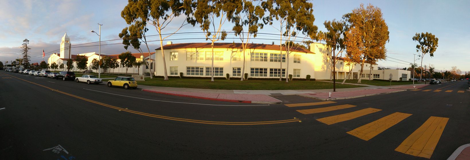 Wide panoramic view of a large white school campus building with trees and a crosswalk in the foreground.