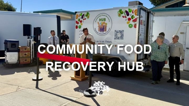 White trailer labeled Community Food Recovery Hub with a speaker at a podium and boxes of produce nearby.