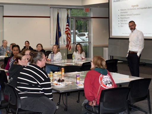 A man in a white shirt presents to a group of adults seated around tables in a meeting room.