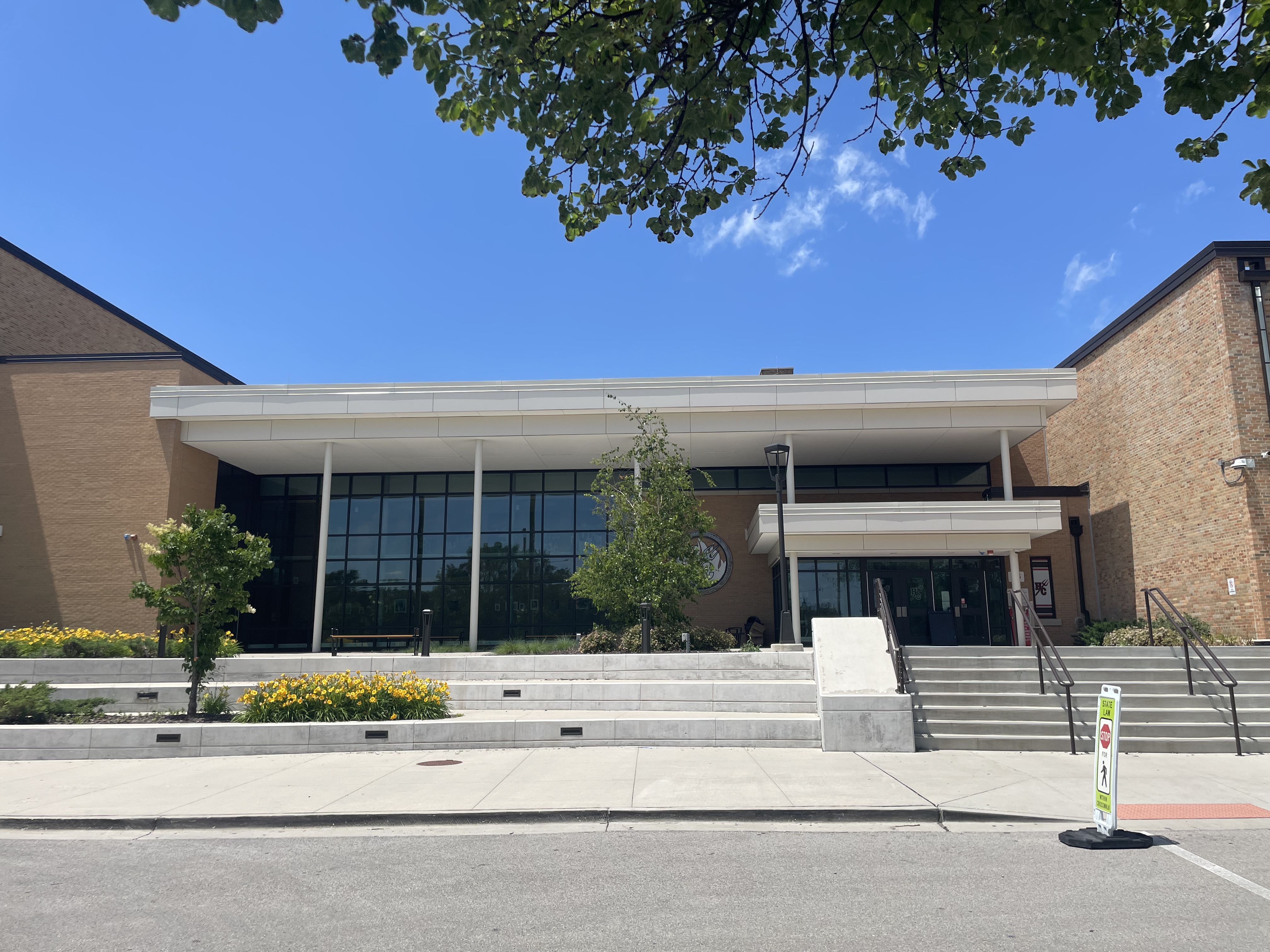Front exterior of a brick and glass high school building with steps, a ramp, and yellow flowers.