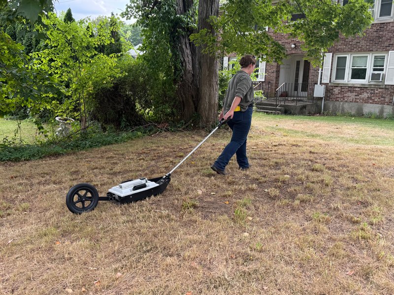 Person pushing a wheeled ground-penetrating radar unit across a dry grassy lawn near a large tree.