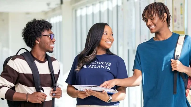 Person in an Achieve Atlanta shirt holds papers while talking with two students in a hallway.