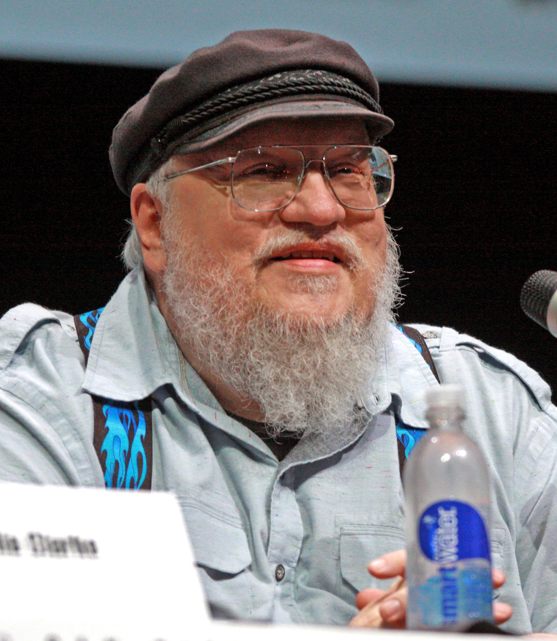 Bearded man in a dark flat cap and glasses seated at a panel table with a microphone.