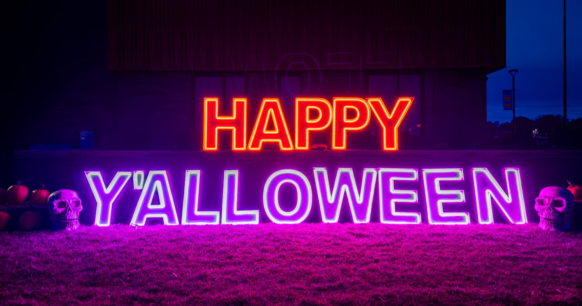 Neon sign reading Happy Y'alloween flanked by glowing skull decorations at night.