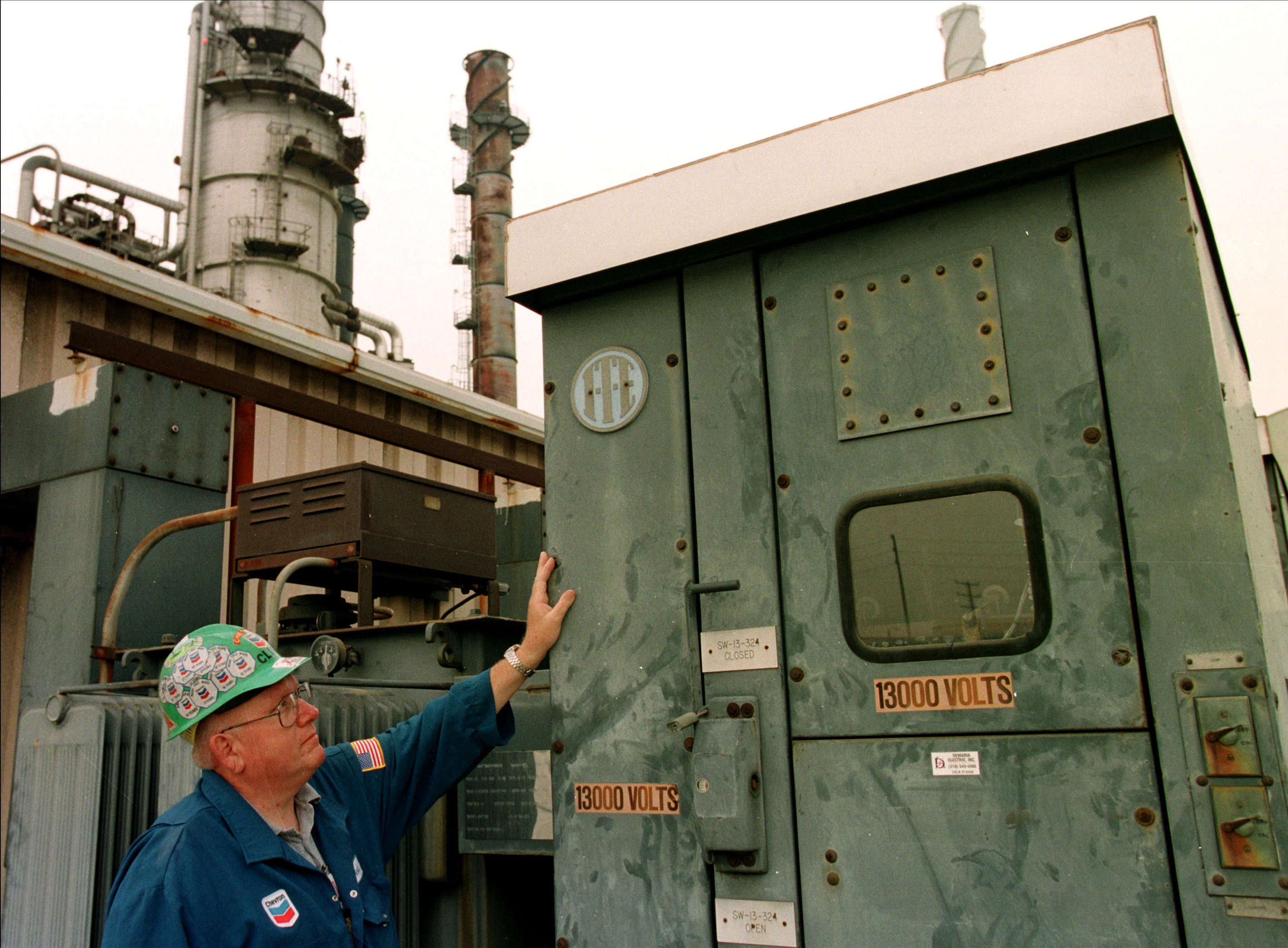 Worker in Chevron hard hat stands next to a large industrial electrical cabinet at a refinery.