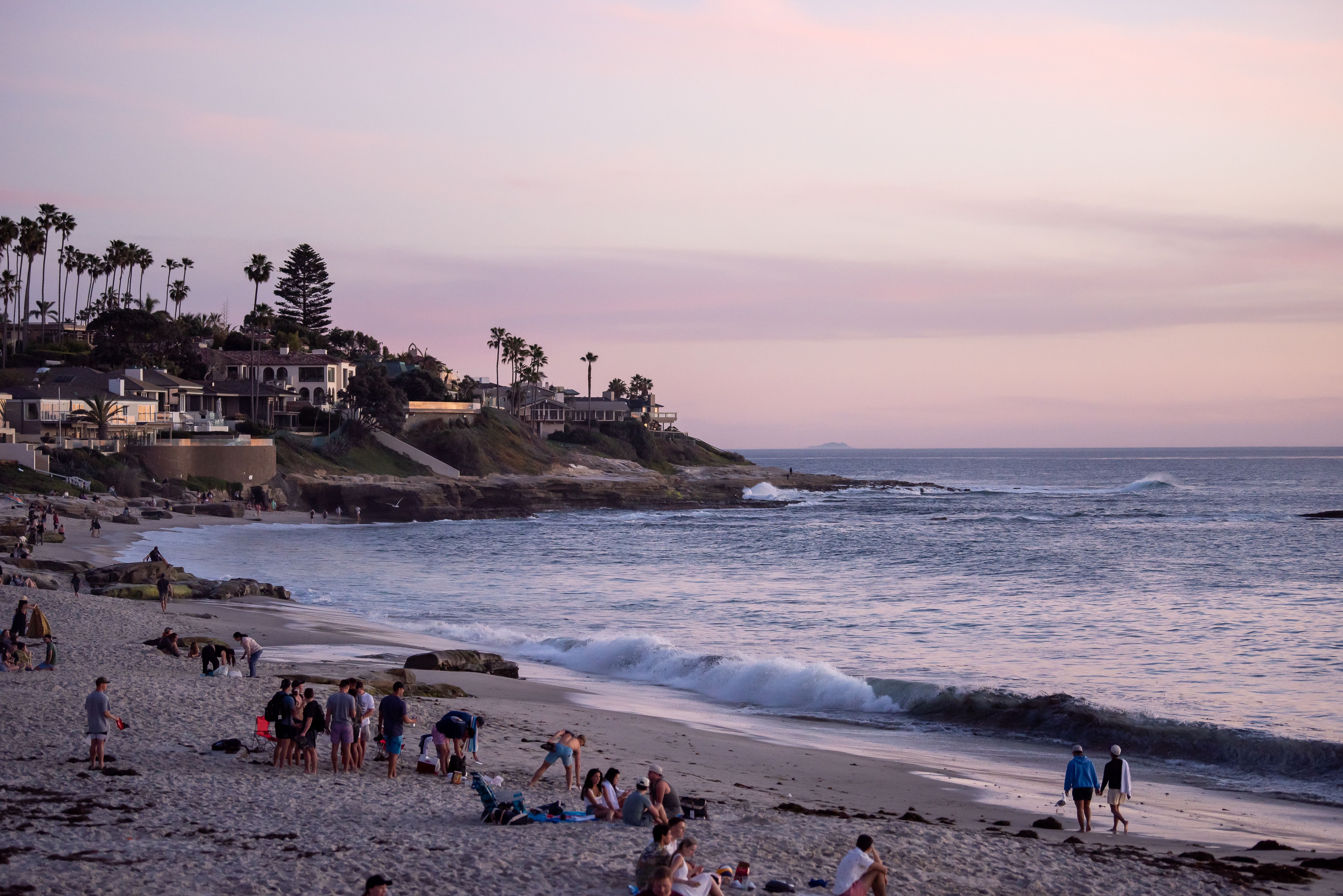 Beachgoers on a sandy shore at sunset with coastal homes on a bluff behind them.
