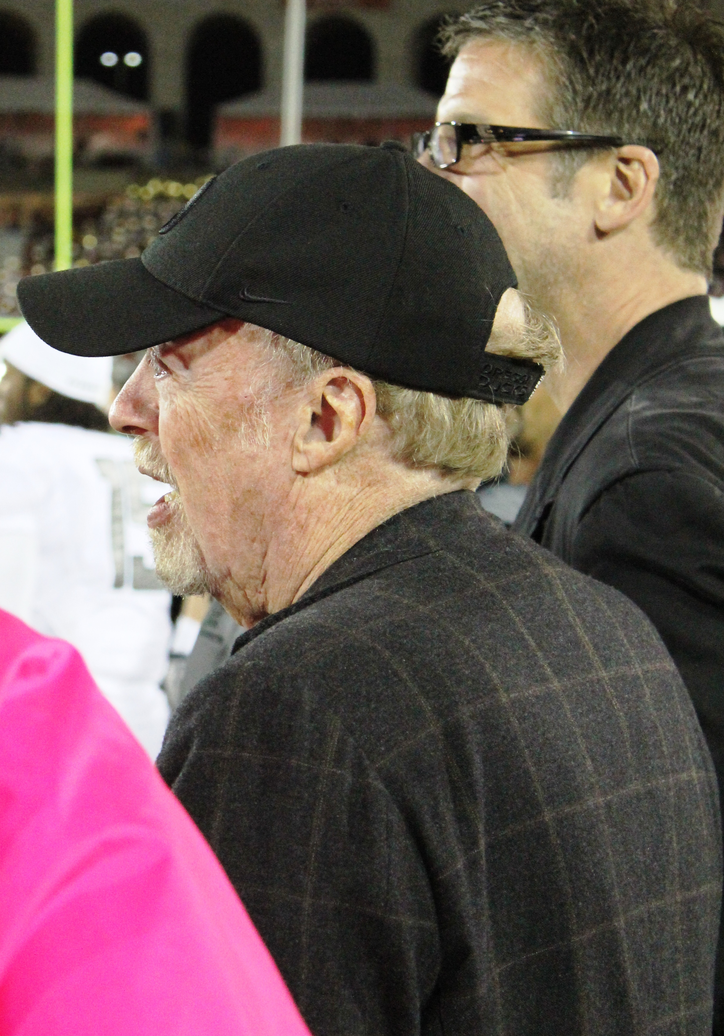 Older man in a black Nike cap and dark plaid jacket at a nighttime sporting event.