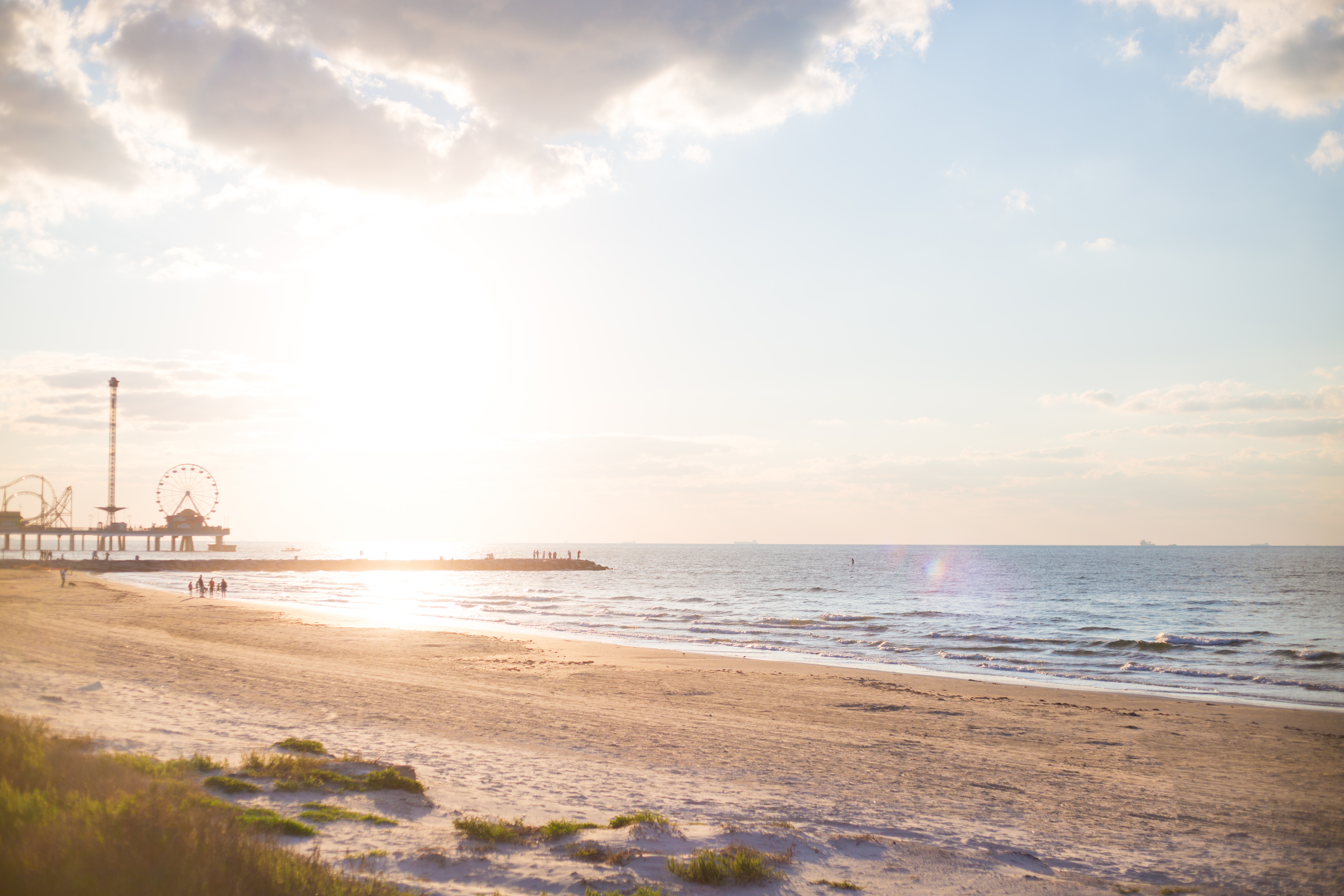 Sandy beach at golden hour with calm surf and a distant pier with a Ferris wheel.