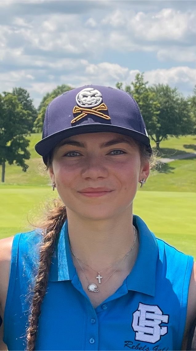 Young female golfer in a blue BC Rebels Golf polo and navy cap on a golf course fairway.