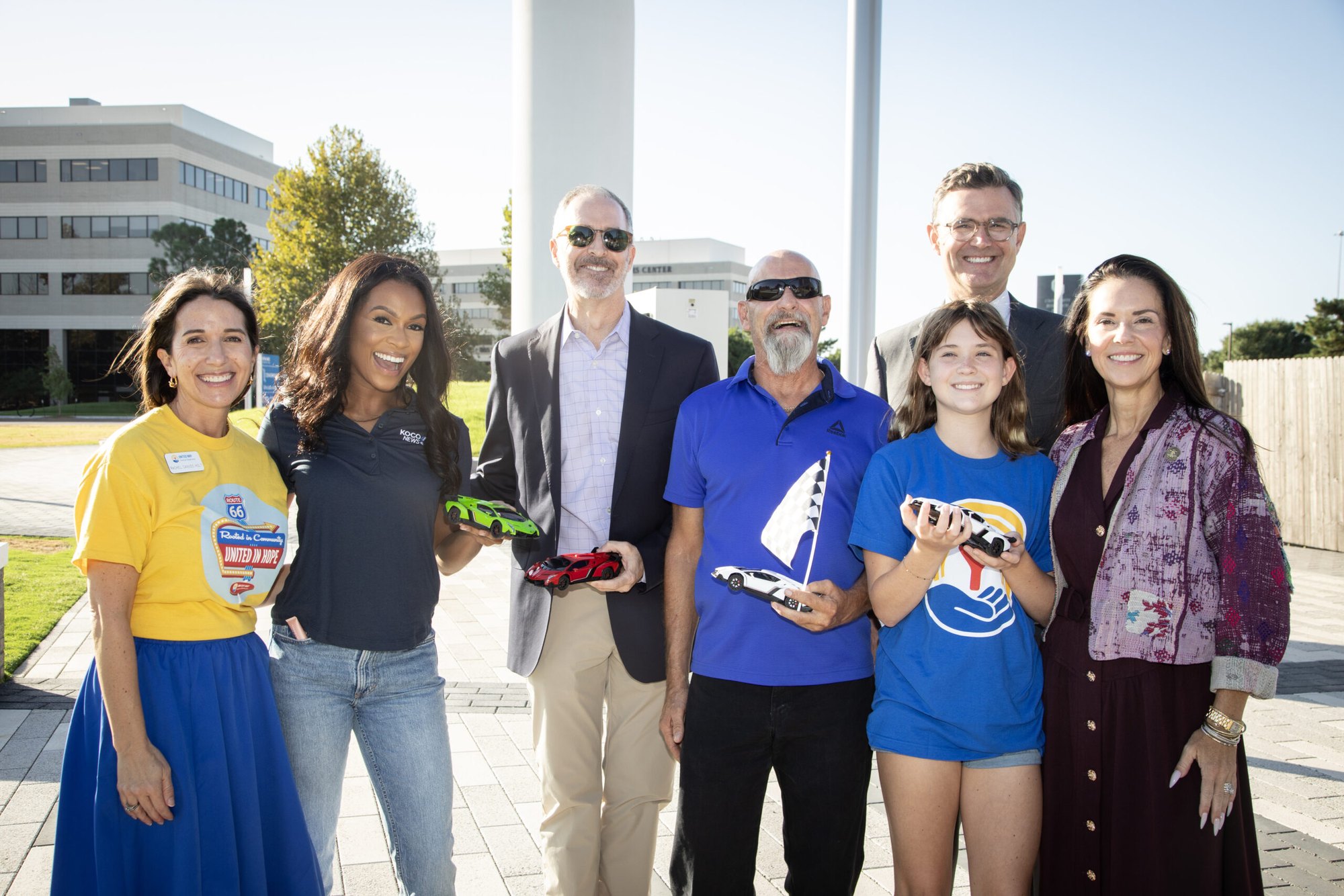 Group of adults and a child posing outdoors holding small remote control cars in front of a modern building.