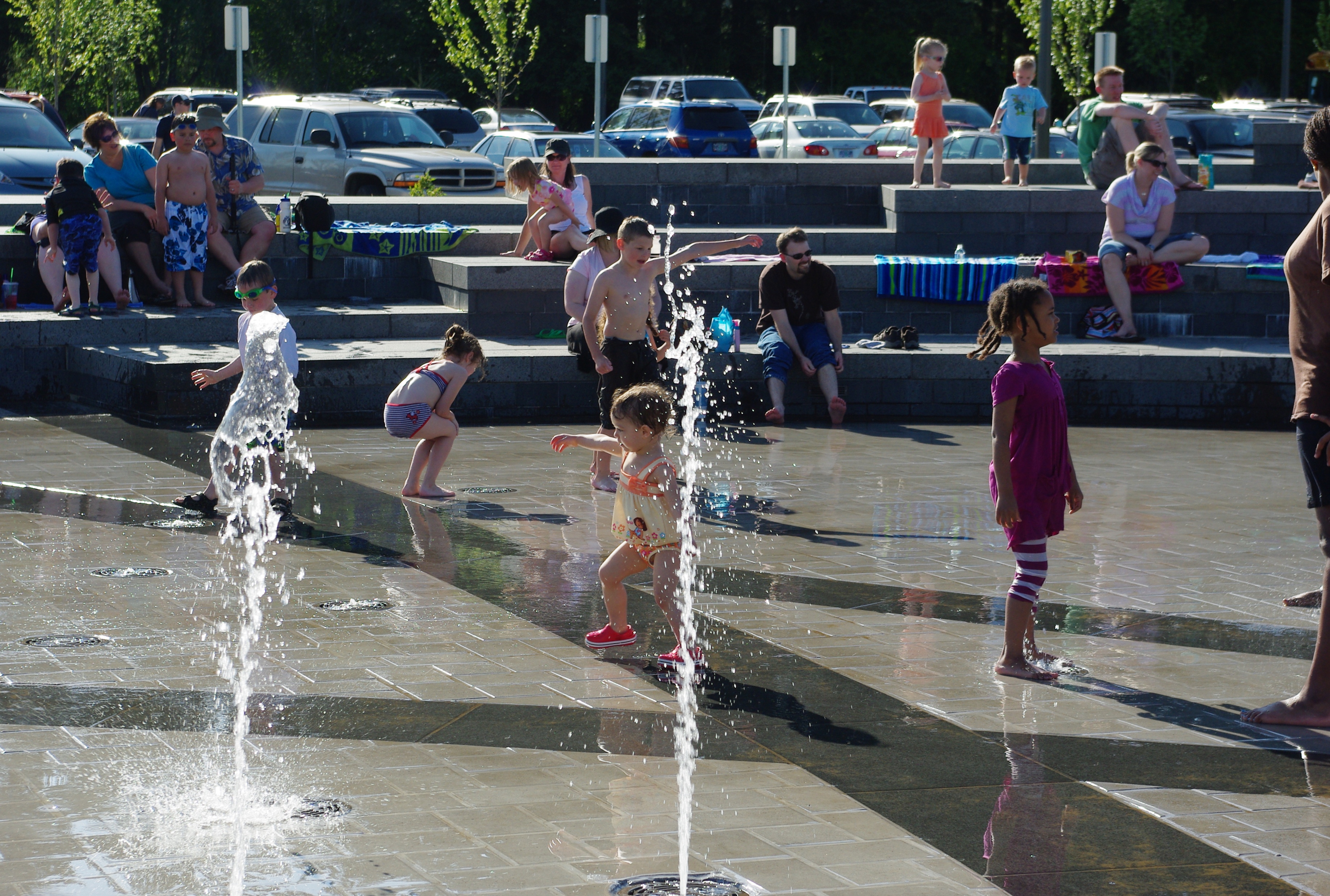 Children playing in ground-level water jets at an outdoor splash pad on a sunny day.