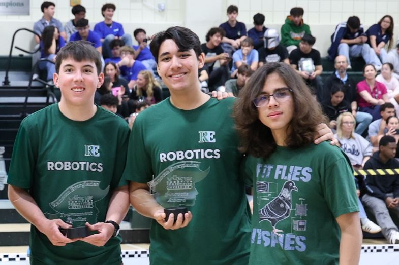 Three students in green robotics shirts hold award plaques inside a gymnasium.