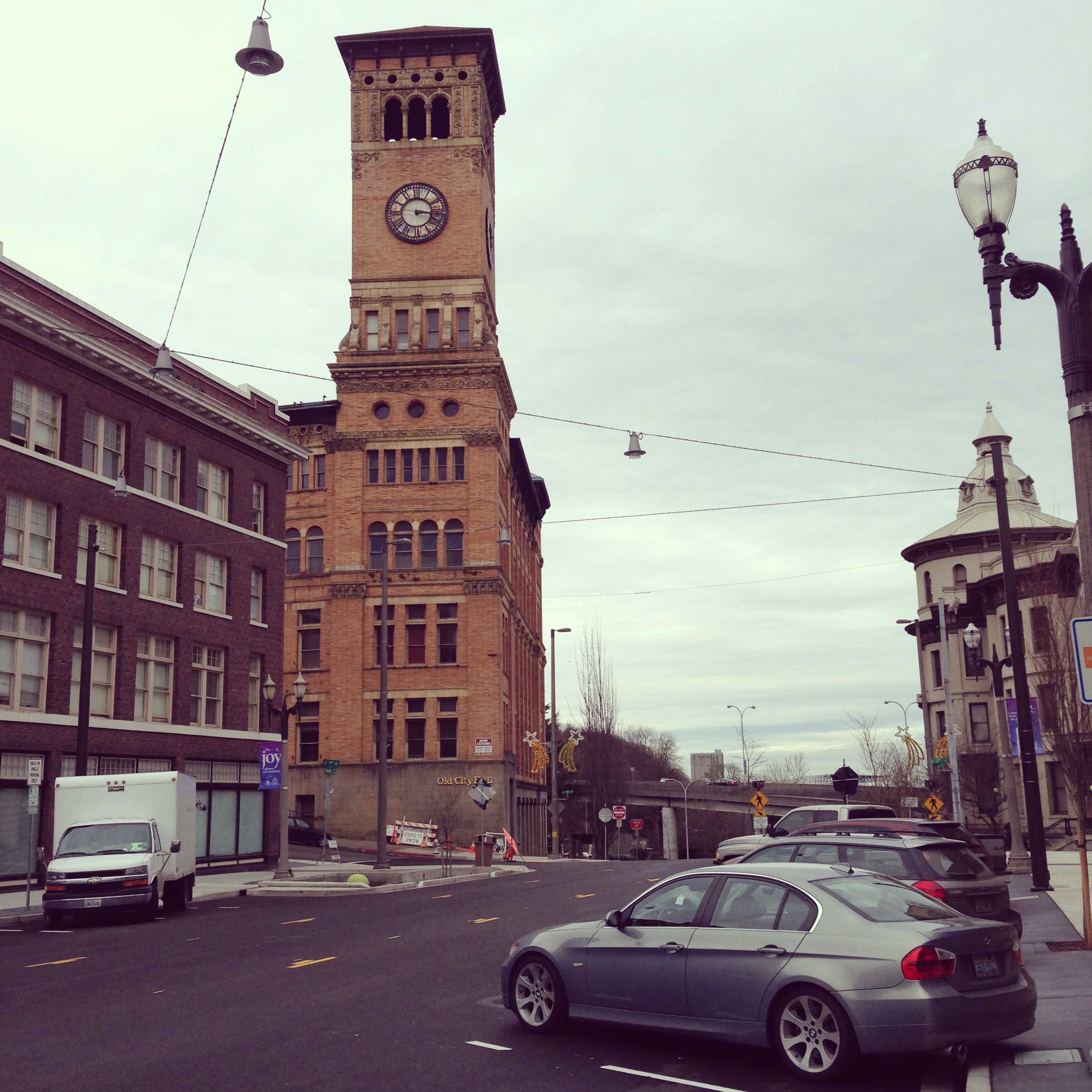 Tall brick clock tower rising above a street corner with cars and lamp posts below.