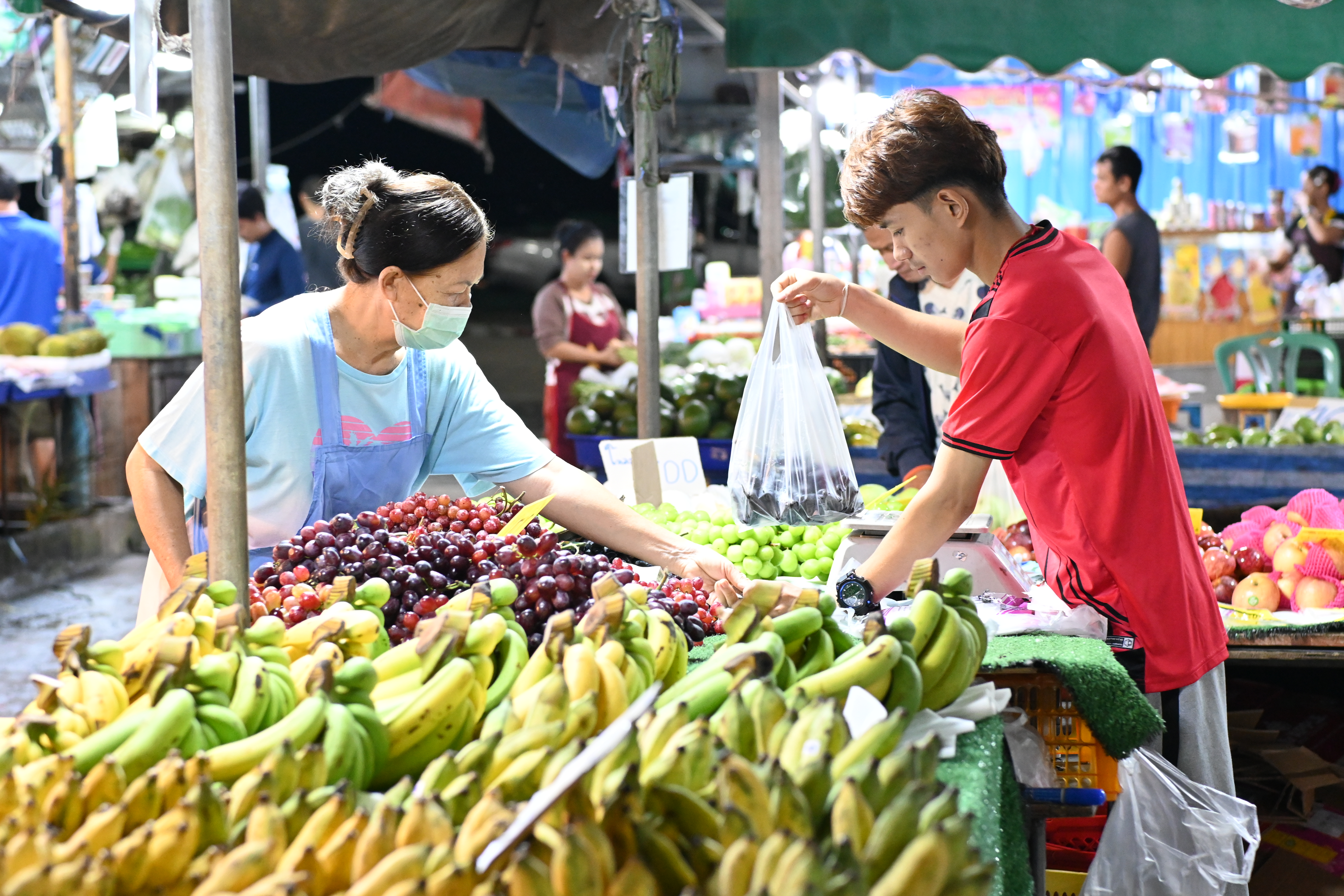 Vendor in blue shirt hands a bag of fruit to a customer at an outdoor market stall.