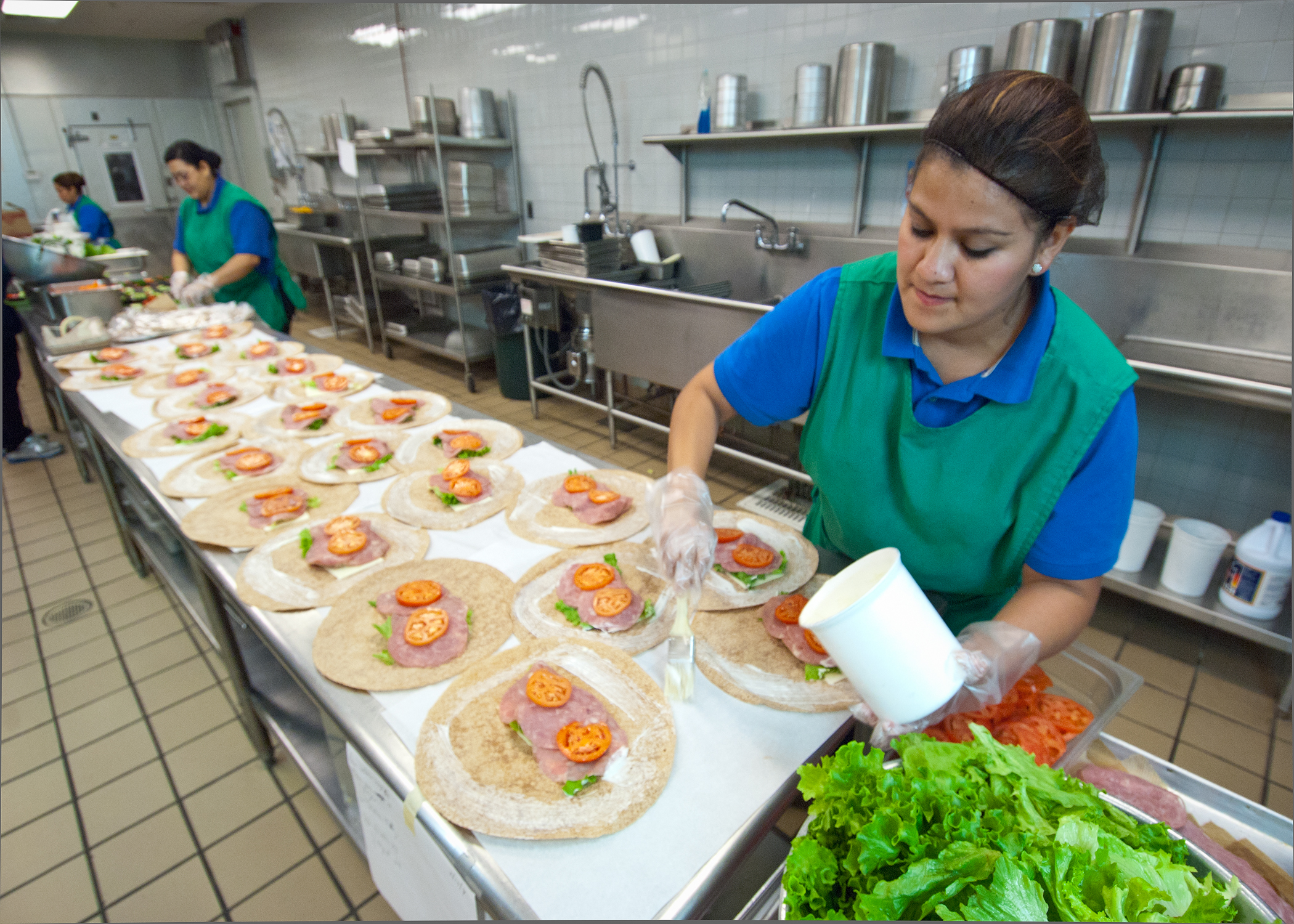 Two workers in aprons and gloves assemble rows of wraps on a stainless steel prep table.