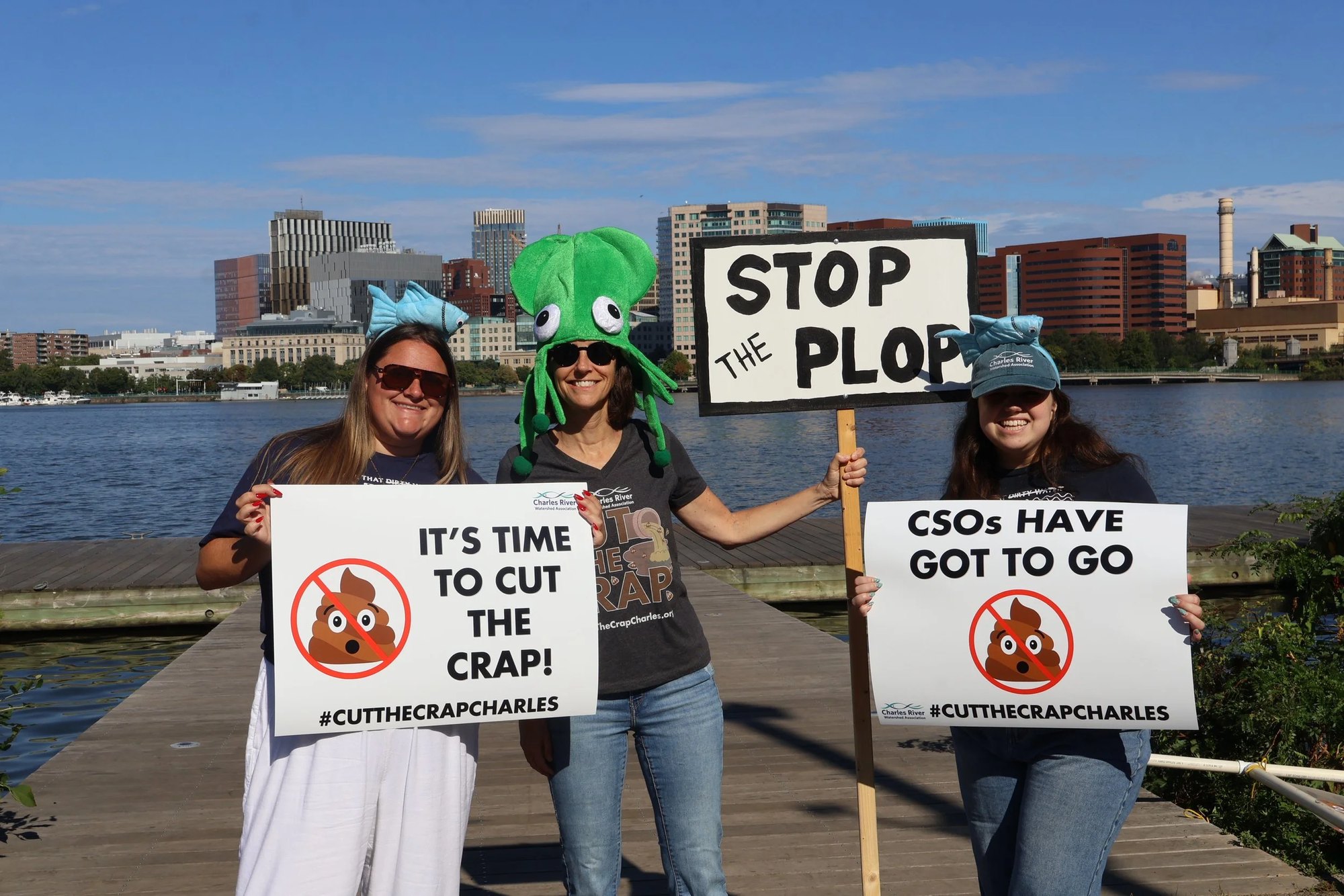 Three women on a dock hold protest signs about stopping sewage overflows into a river.