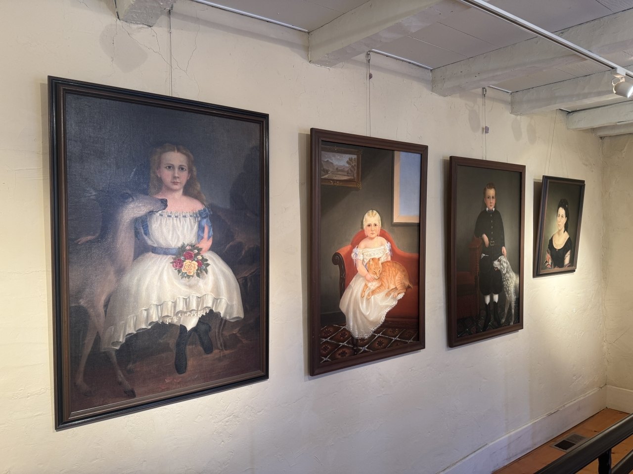 Five framed antique portraits of children and adults hang on a white gallery wall under track lighting.