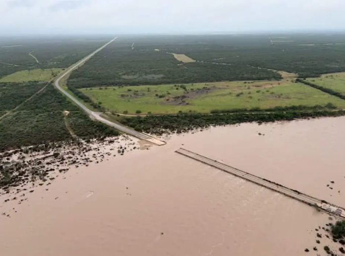 Aerial view of a flooded river swallowing a road amid flat scrubland.
