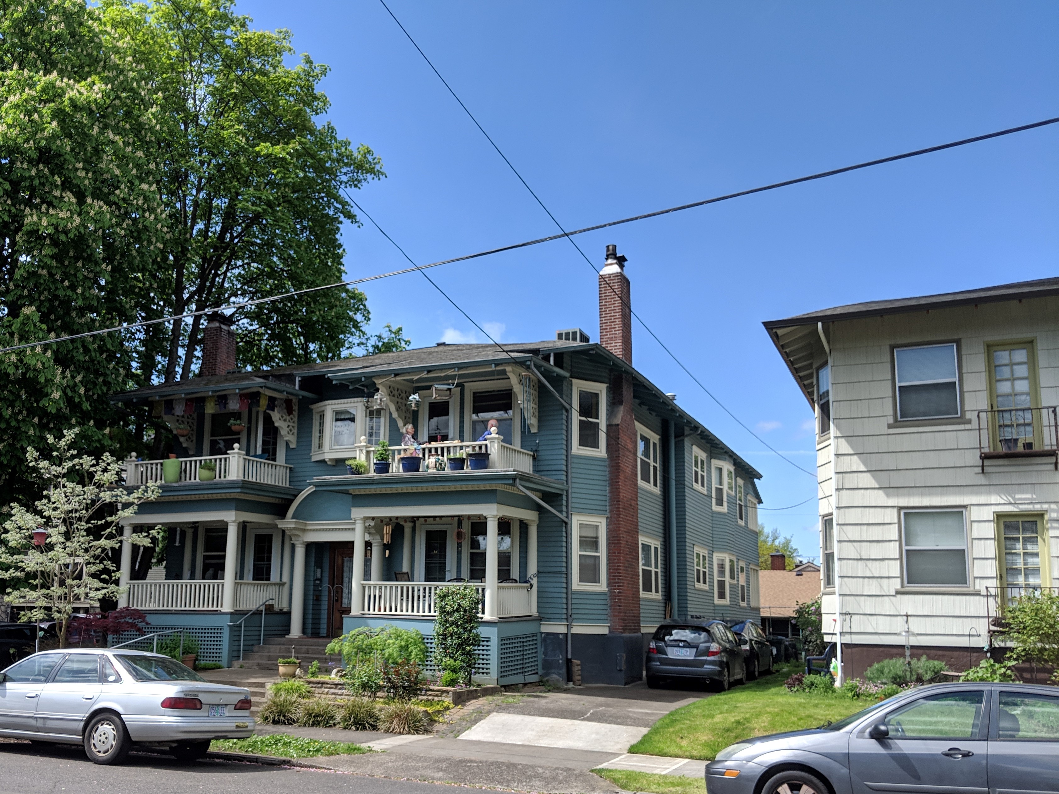 A two-story multi-unit residential building with wrap-around porches on a tree-lined street.