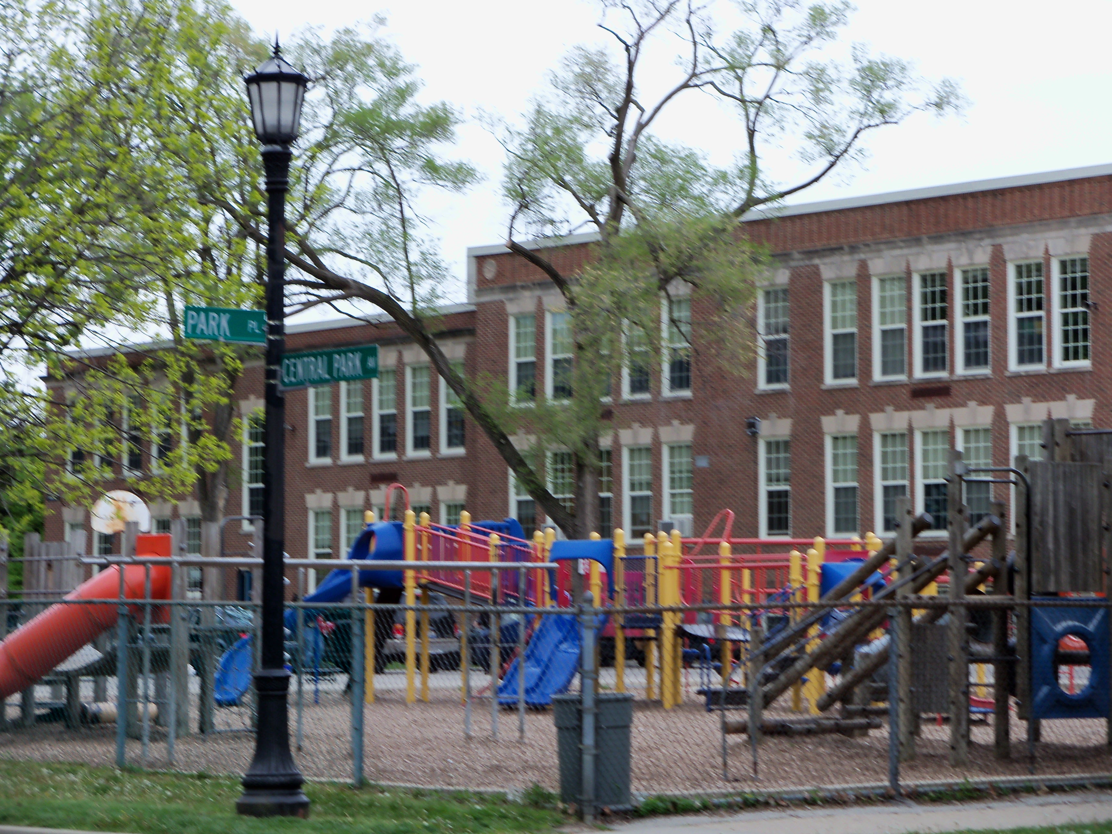 Brick elementary school building with a fenced playground and colorful play equipment out front.