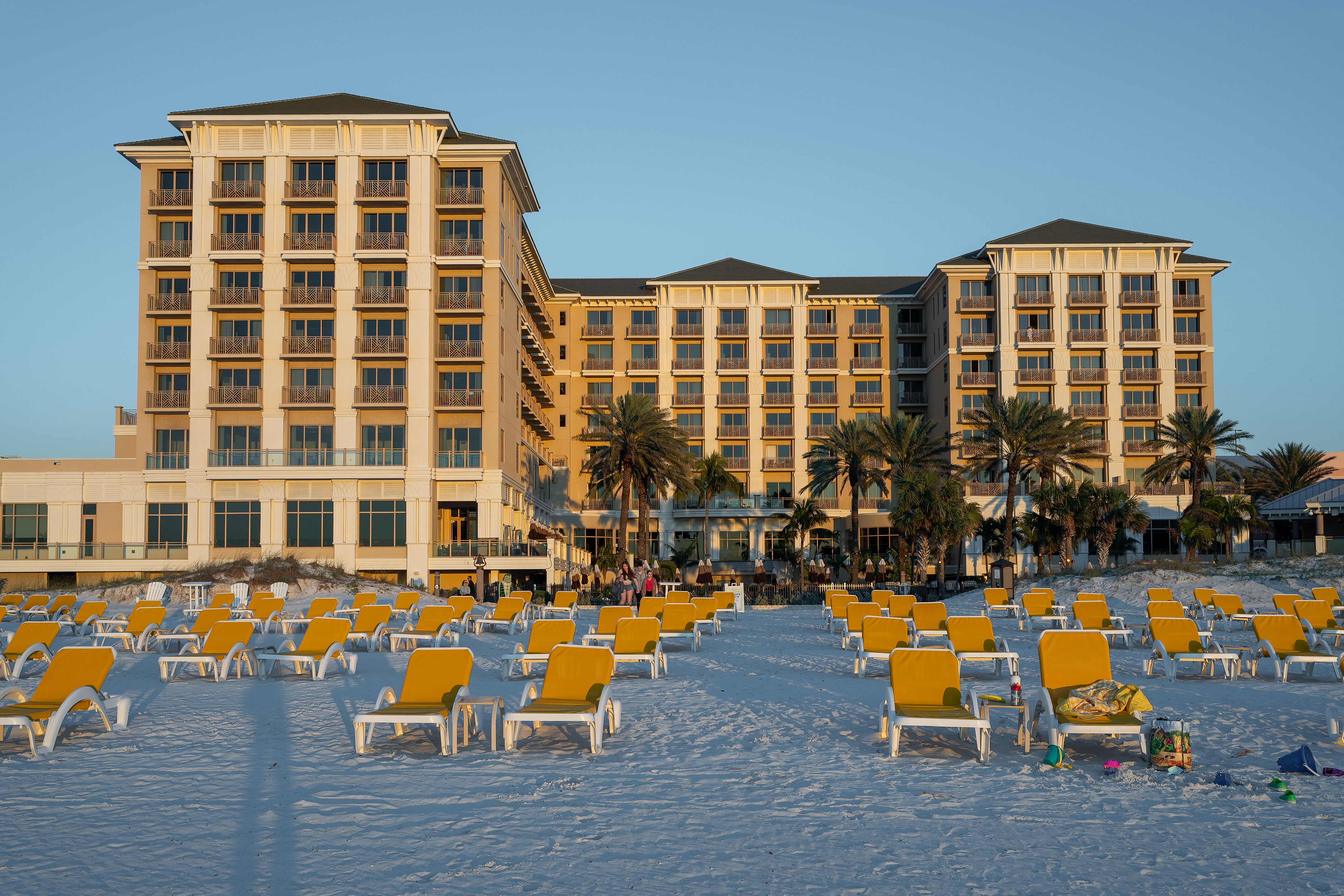 Multi-story beachfront resort hotel with yellow lounge chairs on white sand and palm trees.