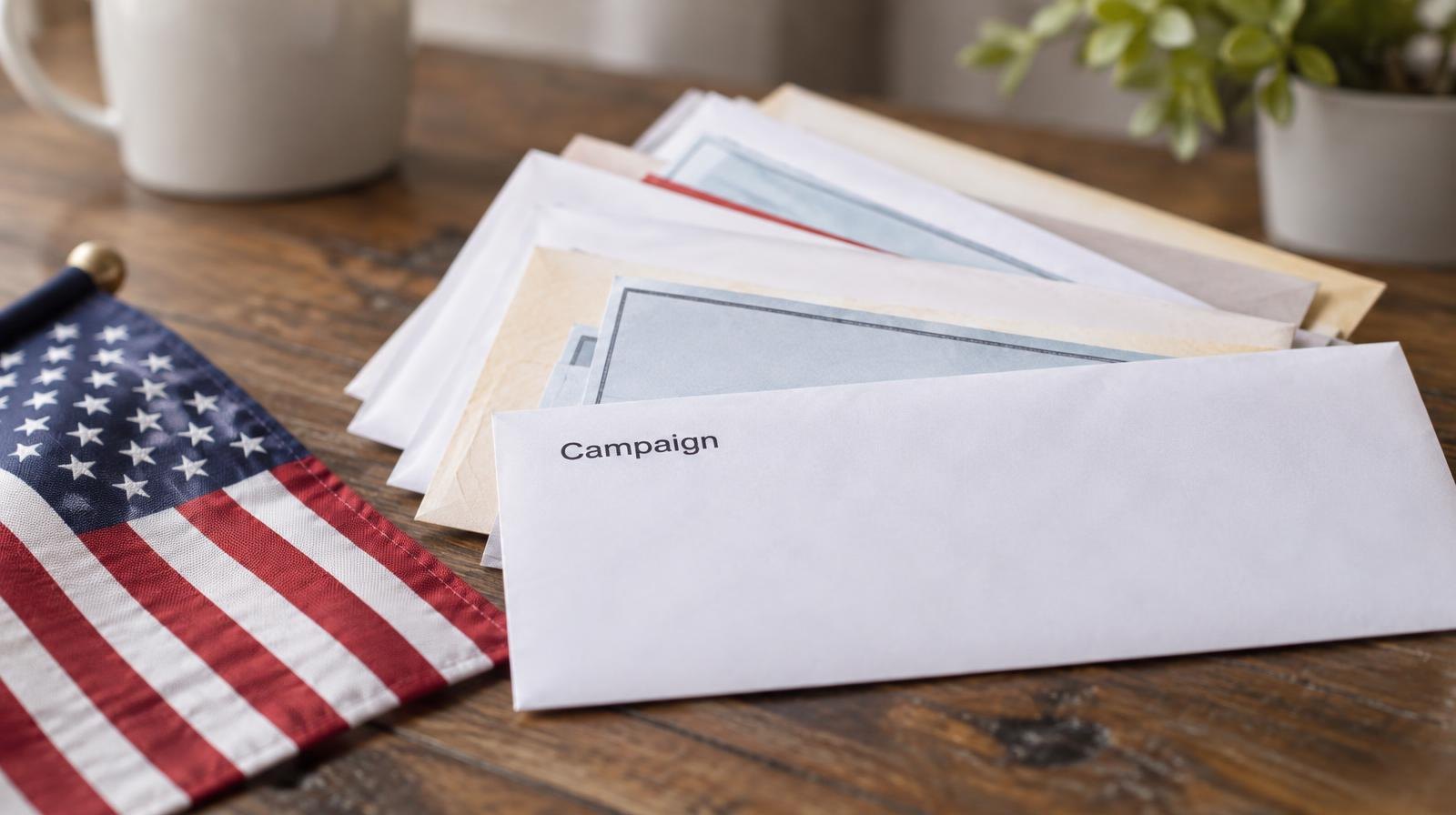 Campaign finance envelopes and checks spread on a wooden surface beside a small flag.
