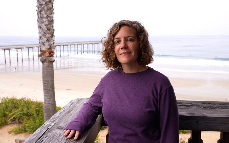 Woman in purple sweatshirt leaning on a wooden railing with a beach and pier behind her.